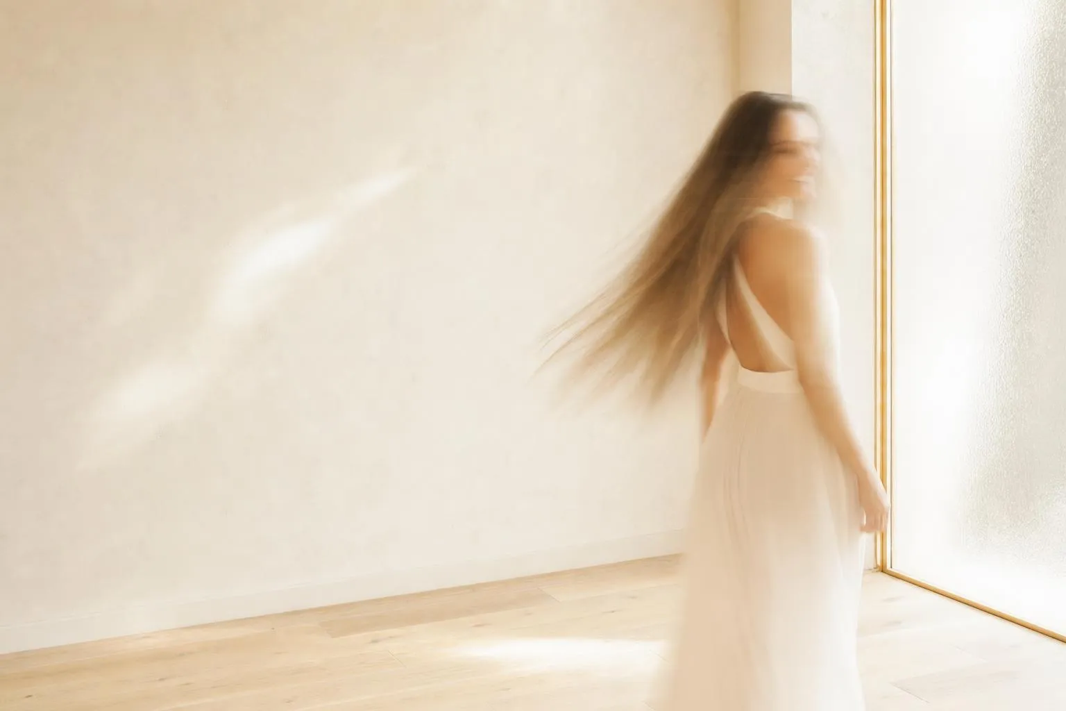 Eine Frau steht in einem hellen, fast leeren Raum am Fenster, in langer Belichtung aufgenommen, sodass ihre Gestalt und ihr langes Haar als weiche Spur im Tageslicht erscheinen.