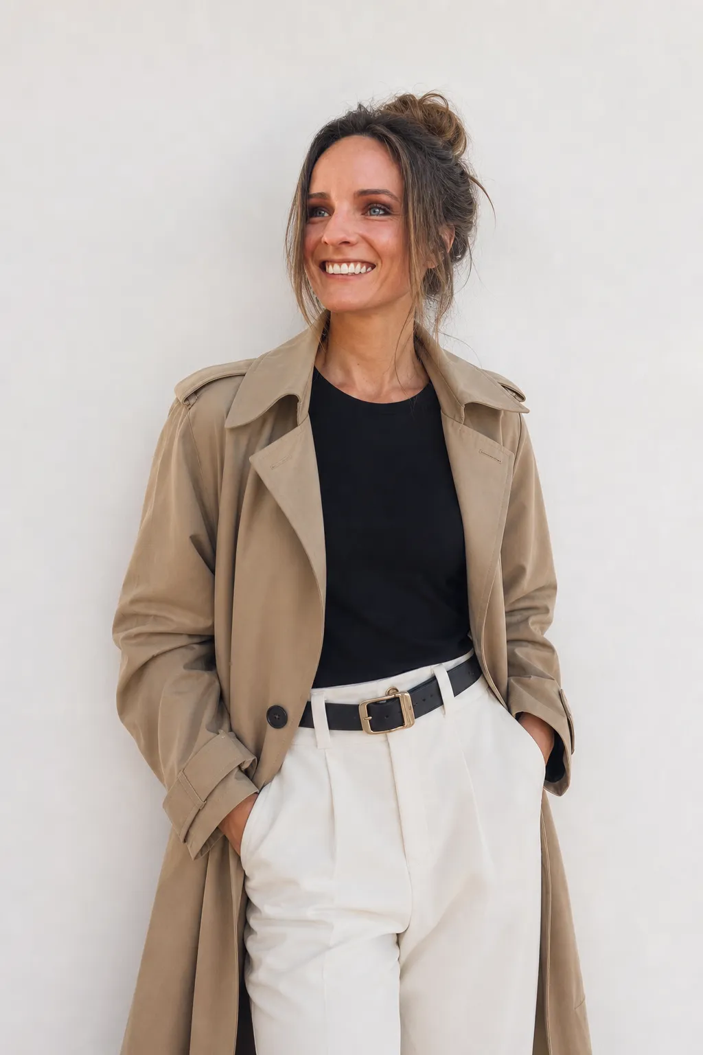 Hannah Steinrock vor einer hellen Wand, in Trenchcoat und weiter Hose, das Haar zu einem lockeren Dutt gesteckt, lachend zur Seite blickend.